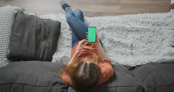 Jovem mulher senta-se em um sofá e começa a usar o telefone em uma tela verde. Tocando na tela, internet online, navegação. Vista superior, dentro de casa, tiro médio. — Fotografia de Stock