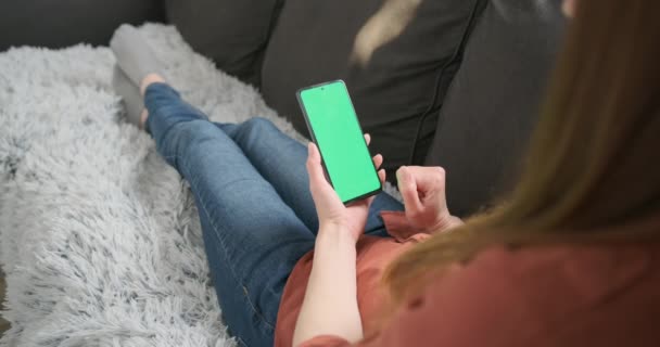 Жінка лежить на дивані, тримає смартфон із зеленим екраном і торкається екрана пальцем. Концепція онлайн-покупки, використовуючи додаток, інтернет-серфінг. Крупним планом . — стокове відео