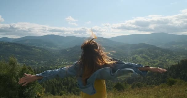 Långhårig flicka, utsträckta armar, står på en kulle med vacker utsikt över de gröna bergen. Vinden blåser, håret svajar, jeansskjortan fladdrar i vinden. Medelhög skott — Stockvideo