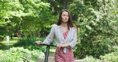 Dişi, yeşil ağaçlı bir parkta elinde elektrikli bir scooter ile geziyor. Çevre kavramı, yeşil enerji, araç. Ön manzara, yakın çekim, portre, gündüz, bahar yazı. Yüksek kalite 4k