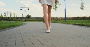 Kadın bacakları parkta yol boyunca yürüyor. Yeşil çimenler, bulutlu gökyüzü. Boş bir park, sakin bir yürüyüş yok, kamera ayakları takip ediyor. Orta boy, ağır çekimde. Yüksek kalite 4k görüntü