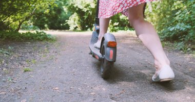 Elbiseli kadın parktaki yolda elektrikli scooterla hareket etmeye başlıyor. Ayağıyla toprağı tekmeliyor. Arka plan, ilkbahar, yaz, gündüz, yeşil ağaçlar yaprakların arasından güneş parlıyor. Yüksek