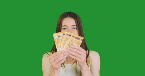一位年轻女子把欧元的钱举到下巴上 轻轻拍着脸的下部 在绿色的背景上眨了眨眼 Chromakey 拥有现金 获得金钱的快乐的概念 — 图库视频影像