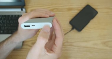 USB kablosunu elektrik bankasına bağlıyorum. Akıllı telefon şarj olmaya başladı. Yakın plan, erkek eli masanın üzerinde dış batarya, telefon ve dizüstü bilgisayar tutuyor. Yüksek kalite 4k görüntü