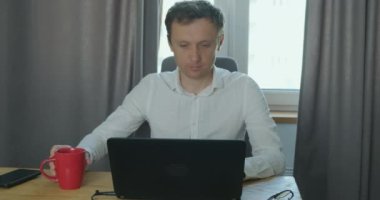 Genç adam dizüstü bilgisayarda çalışıyor ve fincandan kahve içiyor. Hızlandırılmış iş akışı hareketi. Uzaktan kumandalı çalışma kavramı, iş günü, ev ofisi, uzaktan öğrenme, klavyede yazma. Portre, öne.