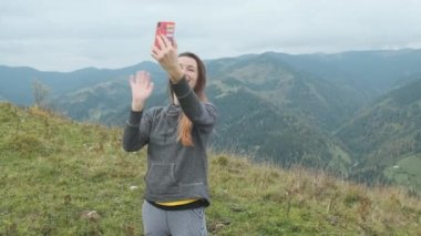 Genç, güzel, uzun saçlı bir kadın Avrupa 'daki dağların tepesinde dururken selfie çekiyor. Saç resimlerini düzeltiyor. Ön manzara, Karpatlar, orta boy, bulutlu, gökyüzü, yeşil dağlar. Yüksek kalite 4k