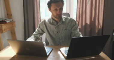 Young IT specialist works on two laptops at the same time. Front view, portrait, home office, desktop. The concept of the modern world, online remote work, how to do everything, overstrain. High