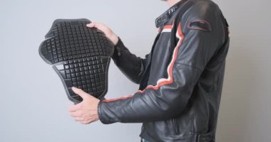 Biker in a leather black sports motorcycle jacket, holds a plastic back protection in his hands, demonstrates it, squeezes it. Side view, close-up, indoors. High quality 4k footage