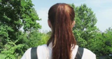 A red-haired girl looks at high-rise residential buildings among green trees and a blue sky. Daytime, summer, city view, back view. High quality 4k footage