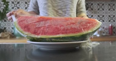 Girl removes a transparent wrapper from a large piece of red juicy watermelon lying on a plate in the kitchen. High quality 4k footage
