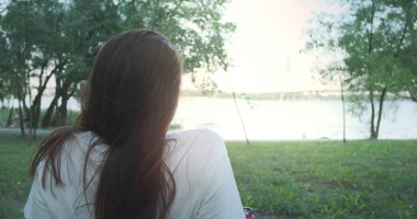 Lonely girl looks at the setting sun in the distance. River, bridge, city park, back view, green grass and trees. High quality 4k footage