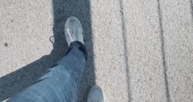 Bir adam asfaltta yürür. Yukarıdan birinci şahıs bakışı. Beyaz spor ayakkabılar, mavi kot pantolon, güneş ışığı ve köprü korkuluğunun gölgesi. Yüksek kalite 4k görüntü