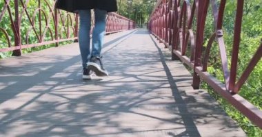 Siyah trençkotlu sırt çantalı yalnız bir kız parkta yaya köprüsü boyunca yürüyor. Arka görüş, geniş açı. Arka planda yeşil yapraklar var. Yüksek kalite 4k görüntü
