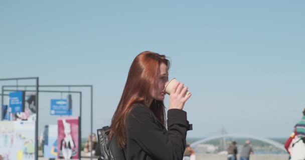 バックパック付きの黒いコートを着た若い女性が紙コップからコーヒーを飲みます 認識できない人々の背景に歩いている ウクライナのキエフの観光客 高品質4K映像 — ストック動画