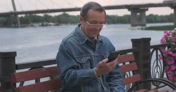 上了年纪戴眼镜的高加索人在智能手机上接电话 河流和大桥在后面 阴云密布 木制长椅和花卉 高质量4K — 图库视频影像