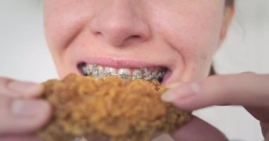 Diş teli takan genç bir kadın fast food yiyor, kızarmış tavuk kanadı yiyor. Yakından, çiğneyen ağız. Abur cuburdan zevk almak, açlığı gidermek. Yüksek kalite 4k görüntü