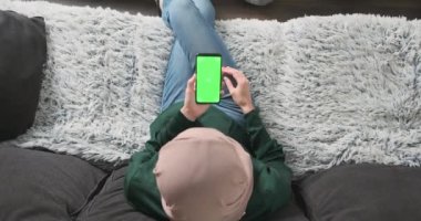 Türbanlı kız elinde yeşil ekranlı akıllı telefon tutuyor. Manzara, oturma odası, kanepe. Uygulamayı kullanarak, yukarı kaydırın. Tanınmayan Müslüman kadın. Yüksek kalite 4k görüntü