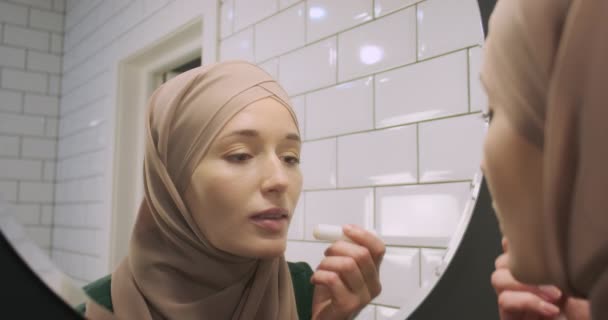 히잡을 아름다운 여인이거울에 접근하여 립스틱으로 입술을 물린다 거울에 초상화 이슬람 — 비디오