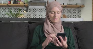 Başörtülü Müslüman kadın oturma odasında kanepede otururken akıllı telefon kullanıyor, portre yapıyor. Mobil uygulama kavramı, öldürme zamanı, online alışveriş, saat içeriği, mesajlaşma. Yüksek kalite 4k görüntü