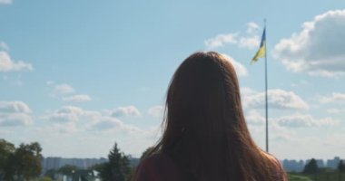 Tanımlanamayan bir kadın rüzgarda sallanan Ukrayna bayrağına bakıyor. Arka manzara, gündüz, beyaz bulutlu mavi gökyüzü. Yüksek kalite 4k görüntü