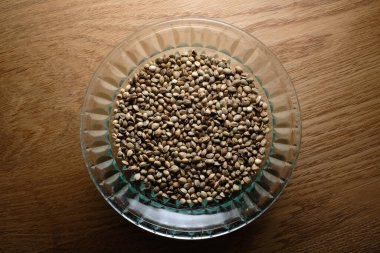 Hemp seeds, marijuana grain in a round glass plate on wooden textured background. Cannabis edible production.
