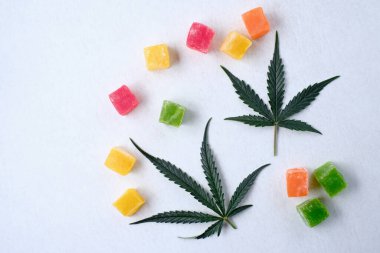 Beyaz dokulu arka planda renkli marmelat ve marihuana bitkisinin yerleşimi. CBD yağlı tatlılar. Üst manzara, düz uzanma.