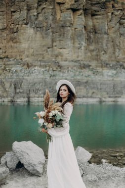 Beyaz gelinlikli güzel bir esmer kız, beyaz şapkalı, bir buket çiçekli, bir turkuaz gölün yanında duruyor dağların arka planında taşların yanında. 