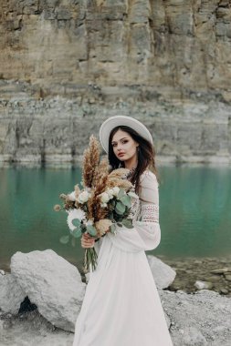 Beyaz gelinlikli güzel bir esmer kız, beyaz şapkalı, bir buket çiçekli, bir turkuaz gölün yanında duruyor dağların arka planında taşların yanında. 