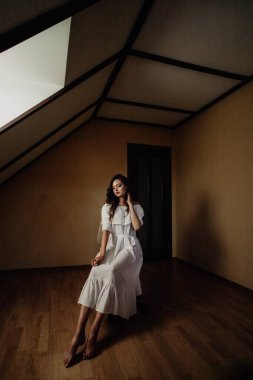 Odada tek başına oturan beyaz elbiseli güzel esmer kız bir sandalyede oturuyor.