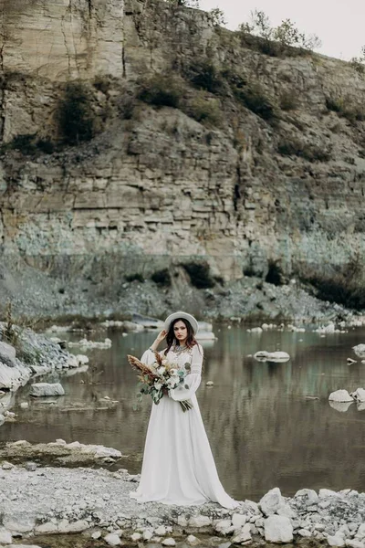 Beyaz bir gelinlik içinde uzun saçlı güzel esmer kızlar, beyaz şapka, boho tarzında bir buket çiçek, yeşil yapraklar, güller ve sazlıklardan oluşan, göl ve dağların yanında duruyor.