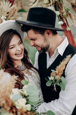 Gelin ve damat yan yana durur ve bir buket çiçekle boho tarzında ve düğün dekorunun yanında gülümserler. Siyah takım elbiseli ve şapkalı Quy ve beyaz elbiseli ve şapkalı bir kız