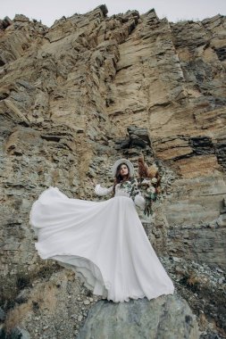 Rüzgarda gelişmekte olan beyaz bir gelinlik içindeki güzel bir esmerin tam boy fotoğrafı, beyaz bir şapka, bir buket çiçekle boho stili bir taş üzerinde taş bir dağ zemininde duruyor.