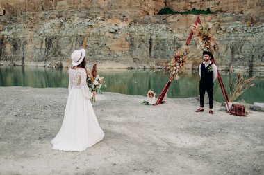 Beyaz gelinlik giymiş bir gelin, boho tarzı bir buket çiçekli bir şapka... damat, düğün dekorunun, gölün, dağların yanında duruyor... ve siyah bir takım elbise, şapka ve beyaz gömlek giyiyor.