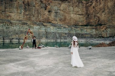 Beyaz gelinlik giymiş bir gelin, boho tarzı bir buket çiçekli bir şapka... damat, düğün dekorunun, gölün, dağların yanında duruyor... ve siyah bir takım elbise, şapka ve beyaz gömlek giyiyor.