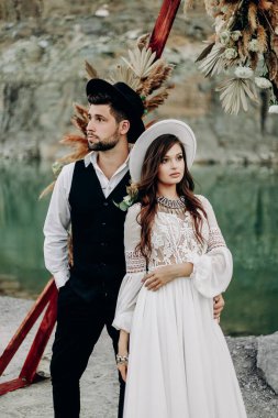 Adam ve kız düğün günlerinde boho tarzı bir düğün dekorunun, dağların, gölün ve taşların yanında dururlar. Beyaz şapkalı bir kız ve siyah takım elbiseli ve beyaz gömlekli bir adam.