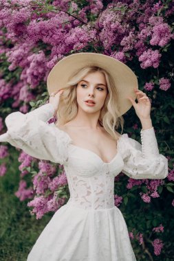 Hasır şapkalı, beyaz gelinlikli, leylak çalılarının yanında açık bluzlu genç sarışın kız.