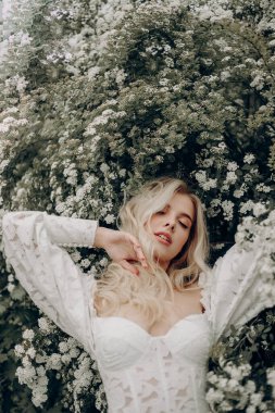 Beyaz gelinlikli, beyaz çiçeklerin yanında açık bluzlu genç sarışın kız.