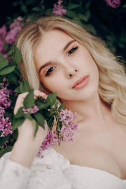 Beyaz gelinlikli, açık bluzlu, leylağın yanında sarışın bir kız.
