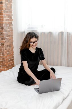 Good morning concept. Beautiful sexy young brunette girl with glasses wearing a black velvet pajama sleepwear on white bed in light cozy bedroom with laptop