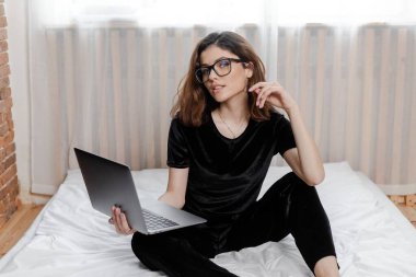 Good morning concept. Beautiful sexy young brunette girl with glasses wearing a black velvet pajama sleepwear on white bed in light cozy bedroom with laptop