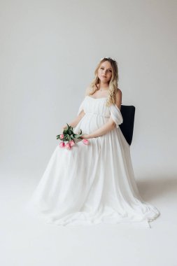 Beyaz elbiseli, sarışın hamile bir kadın. Beyaz bir arka planda. Elinde bir buket lale çiçeği var.
