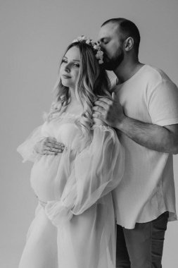 Beyaz elbiseli, beyaz arka planda, kafalarında çelenk olan, elleri karnında, beyaz elbiseli, hamile ve sarışın bir kadın.