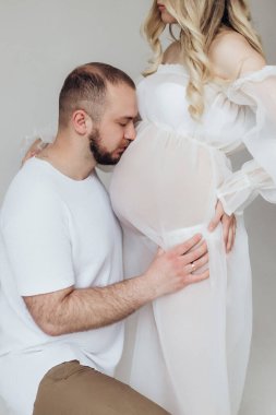 Beyaz şeffaf elbiseli, beyaz arka planda, kafalarında çelenk olan ve ellerini karnında tutan sarışın koca ve hamile eş.