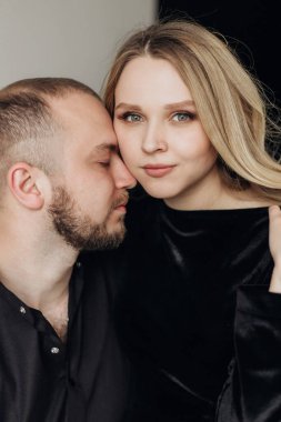 Siyah arka planda siyah elbiseli hamile bir kadın ve koca bir sandalyede oturup sarılıyor.