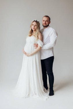 Beyaz elbiseli, beyaz arka planda, kafalarında çelenk olan, elleri karnında, beyaz elbiseli, hamile ve sarışın bir kadın.