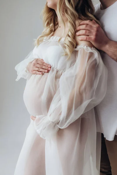 夫と妊娠中のブロンド妻でAホワイトドレスでスタジオの上に白い背景で彼らの頭の上に花輪で彼らの手を保持彼らの胃 — ストック写真