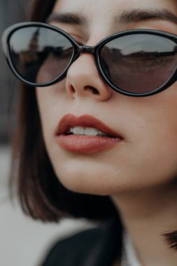 Siyah ceketli, siyah gözlüklü esmer bir kız şehirde el çantasıyla oturuyor.