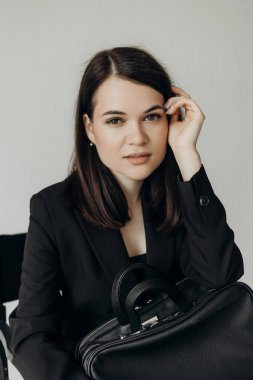A girl in a black suit with a bag on a white background sits on a chair