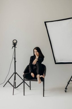 A girl in a black suit on a white background sits on a chair