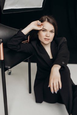 A girl in a black suit on a white background sits on the floor near a chair